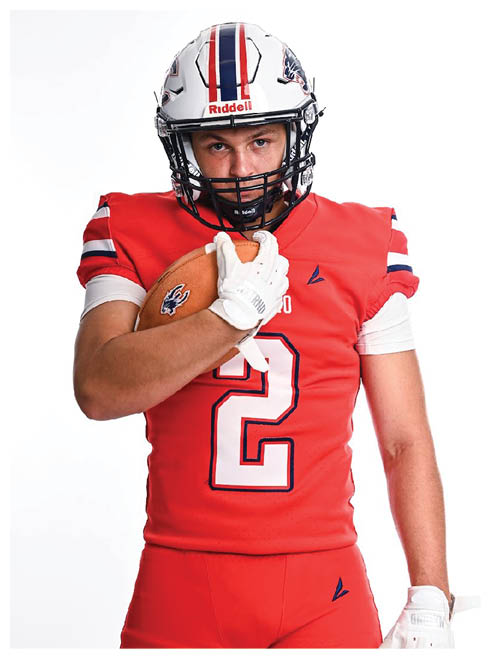 A person wearing a red football uniform and white helmet is holding a football close to their chest while looking forward. Description generated by AI