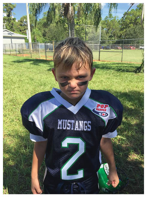 A child wearing a Mustangs football jersey with the number 2 and black face paint stands on grass holding a green helmet, looking serious. Description generated by AI