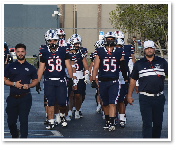 A group of people wearing football uniforms and helmets walk together outdoors accompanied by two people in matching polo shirts and caps. Description generated by AI