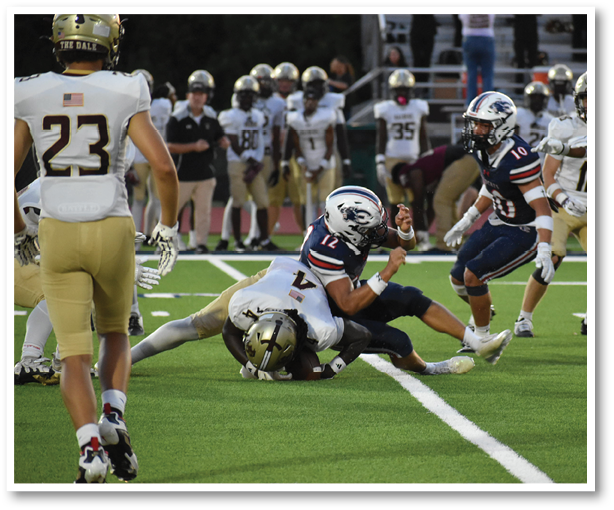 A group of people wearing football uniforms are engaged in a tackle on a green field with white yard lines. Description generated by AI