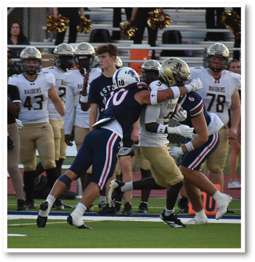 A group of people stands on the sidelines of a football field watching a player in a navy blue uniform with the number 10 tackle or block a player in a white and gold uniform. Description generated by AI