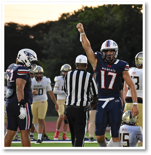 A person in a Wildcats football uniform raises one arm while standing near a referee and other players on a football field during a game. Description generated by AI
