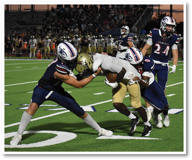 Three people in football uniforms are engaged in a tackle on a green field during a game. Description generated by AI