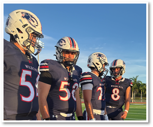 Four people wearing Wildcats football uniforms and helmets stand outdoors under a clear blue sky. Description generated by AI