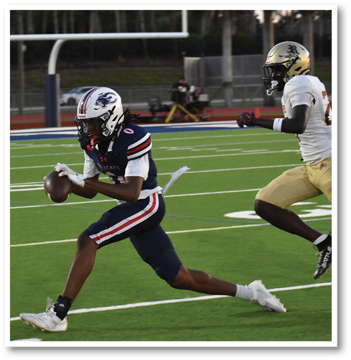A person in a dark blue football uniform is running with the ball while another person in a white and gold uniform chases them on a football field. Description generated by AI