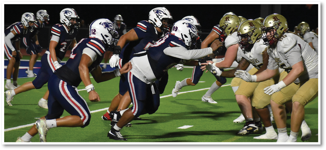 Two opposing American football teams in helmets and uniforms are lined up facing each other on a field at dusk, preparing for a play. Description generated by AI