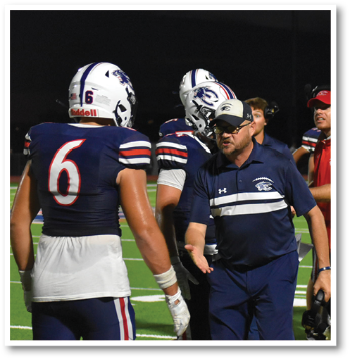 People in sports uniforms are standing and walking on a football field with a coach in a navy blue outfit giving instructions. Description generated by AI