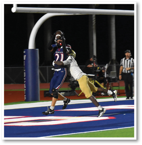 A person in a dark sports uniform catches a football in the end zone while two people in light uniforms attempt to defend during a nighttime football game. Description generated by AI