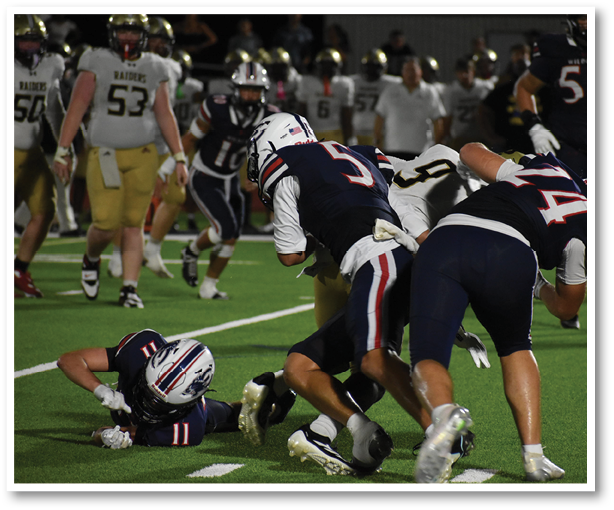 Several people wearing football uniforms are engaged in a play on a green field with one person on the ground and others bending or running nearby. Description generated by AI