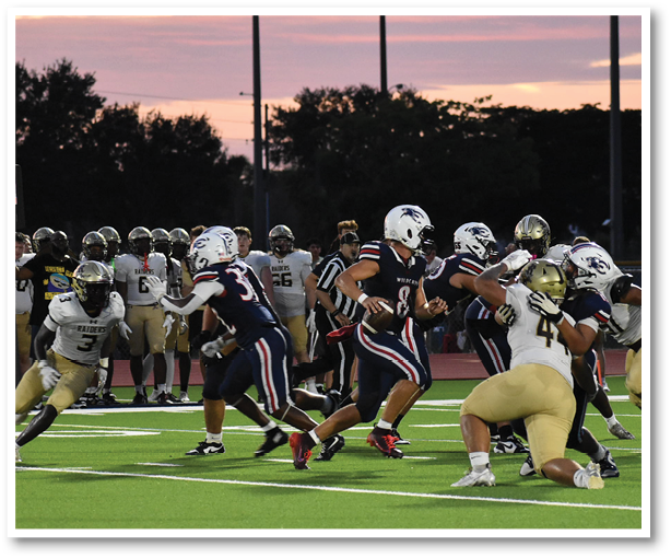 A group of people wearing football uniforms are actively playing on a green field during sunset with spectators and team members visible in the background. Description generated by AI