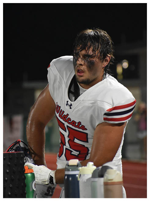 A person with wet hair and black face paint under their eyes is wearing a white sports jersey and appears focused or tired against a dark background. Description generated by AI