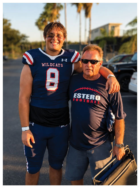 A person in a Wildcats football uniform stands with an arm around another person wearing sunglasses and an Estero Football shirt while holding a folded chair in a sunny outdoor parking area with palm trees in the background. Description generated by AI