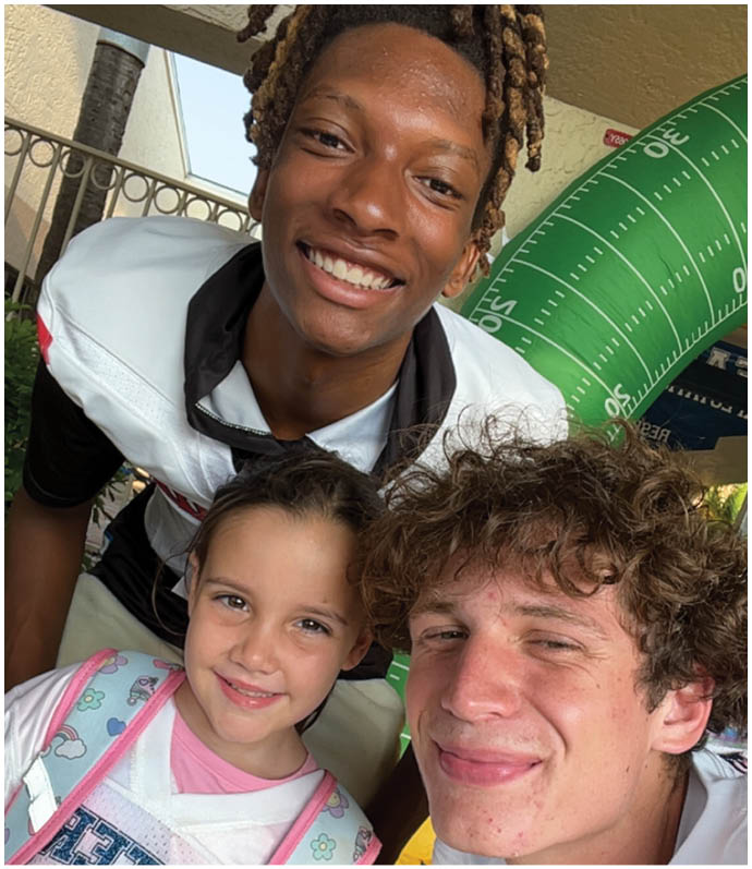 Three smiling people with different hairstyles pose closely together in a casual outdoor setting with a large inflatable football decoration in the background. Description generated by AI