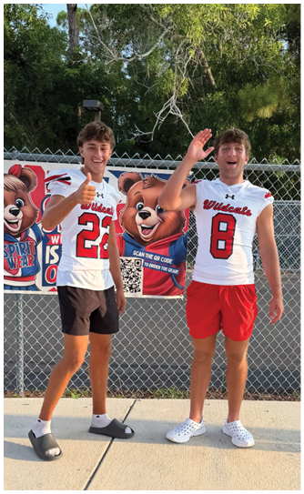 A person wearing a white sports jersey with the number 22 is standing and giving a thumbs-up in front of a chain-link fence with colorful cartoon bear mascots on banners. Description generated by AI