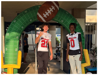Two people wearing football jerseys stand under a large inflatable arch shaped like a football and a green field with yard lines. Description generated by AI