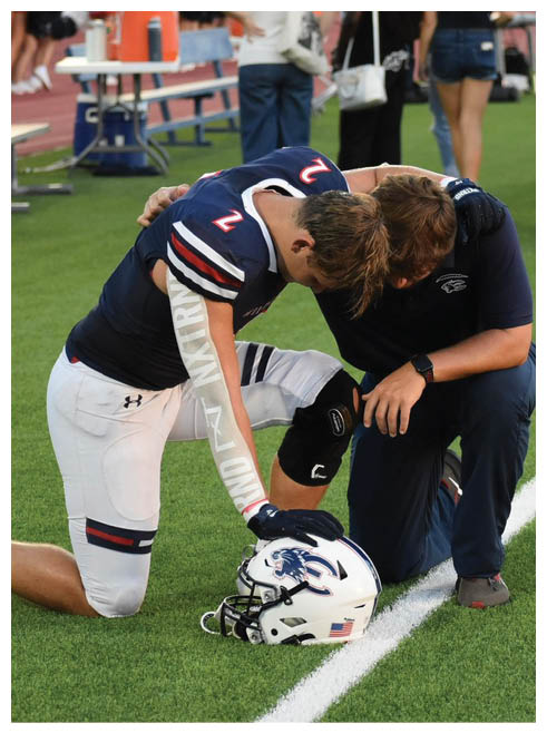 An adult wearing a football uniform with the number 2 is bent over on a field while another person supports them with a hand on their back. Description generated by AI