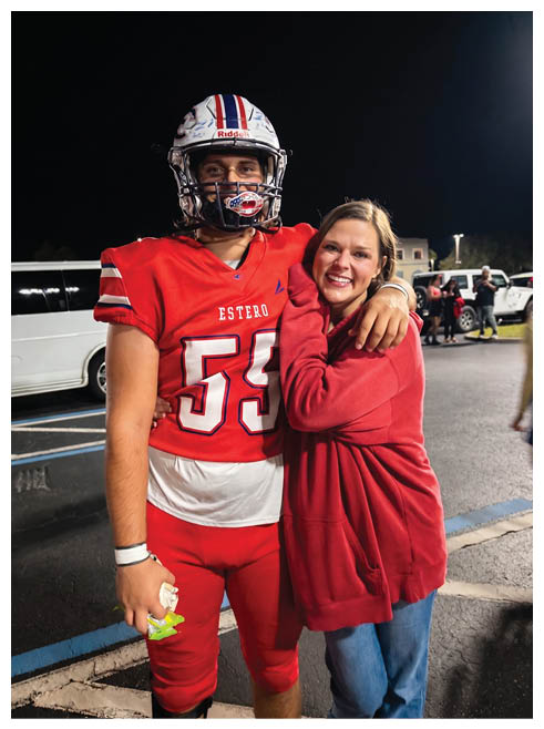 A person wearing a red football uniform and helmet stands next to another person in a red sweatshirt who is smiling and leaning on their shoulder against a dark background. Description generated by AI
