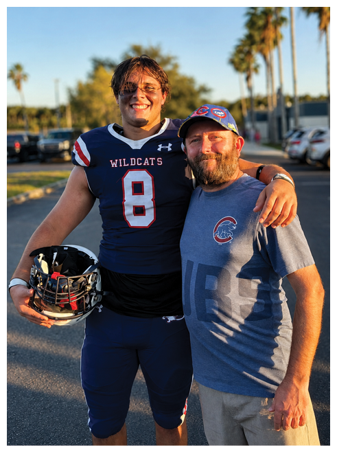 A person in a Wildcats football uniform holding a helmet stands next to another person wearing a Cubs baseball cap and t-shirt, both smiling outdoors with palm trees and parked cars in the background. Description generated by AI