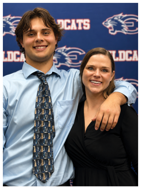 Two smiling adults stand close together with one person’s arm around the other in front of a blue backdrop with text and a wildcat logo. Description generated by AI