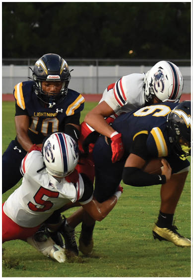 Two people in football uniforms are engaged in a tackle on a grassy field with trees and palm trees in the background. Description generated by AI