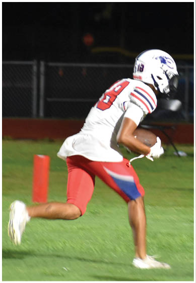 A person wearing a white football uniform with red and blue accents is running on a grassy field while holding a football. Description generated by AI