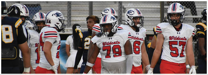 People wearing football uniforms are standing on a field, some holding helmets, with a chain-link fence and bleachers in the background. Description generated by AI