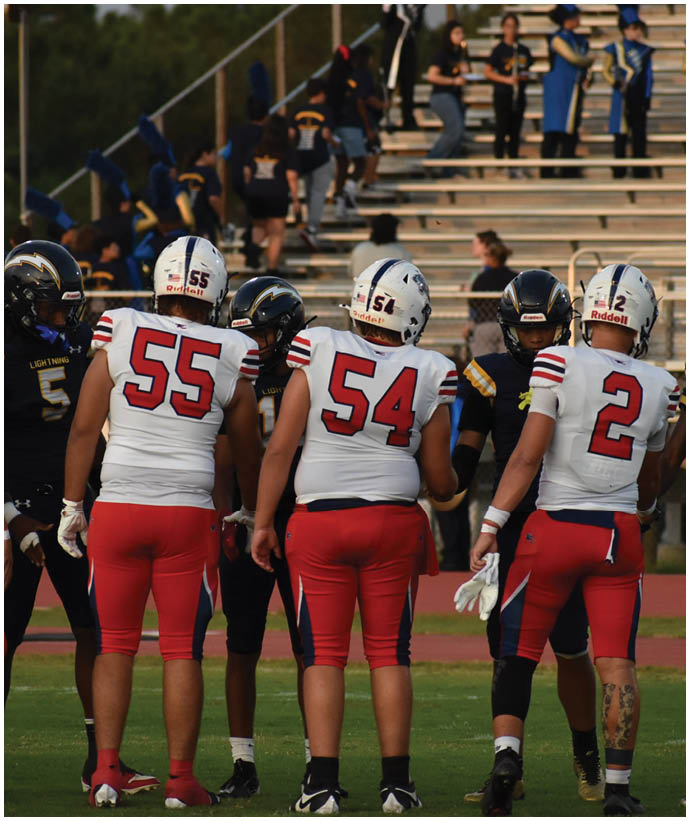 Three people in white and red football uniforms with helmets stand on a field facing away from the camera while others in dark uniforms stand nearby. Description generated by AI