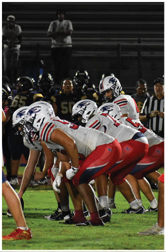 Adults in football uniforms are positioned on a field preparing for a play during a nighttime game. Description generated by AI