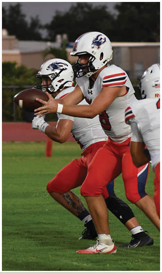 Two people wearing white football helmets and uniforms are engaged in a play with one person handing off a football to the other on a field with trees and buildings in the background. Description generated by AI