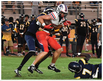 A person in a red and white football uniform is running with the ball while three people in dark blue and yellow uniforms attempt to tackle them on a grassy field with spectators in the background. Description generated by AI