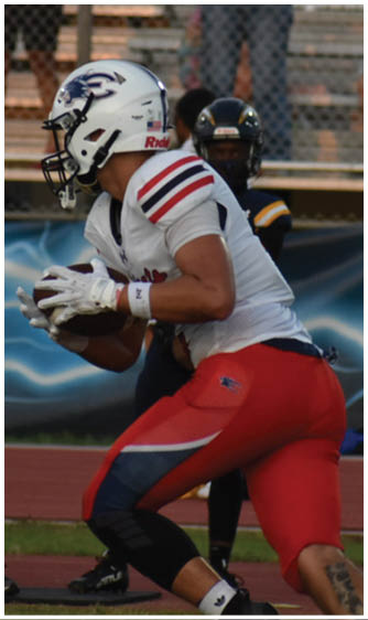 A person in a white football uniform with red and blue accents is holding a football and preparing to run while another person in a dark uniform is behind them on a sports field with spectators in the background. Description generated by AI