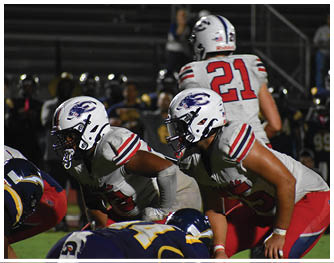 Two people in white and red football uniforms are crouched and ready to play against opponents in dark blue and yellow uniforms on a football field at night. Description generated by AI