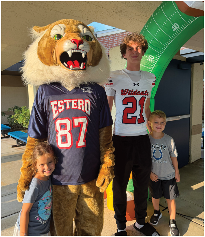 Three children stand smiling next to a person in a large wildcat mascot costume wearing a sports jersey, with an inflatable football goalpost in the background. Description generated by AI