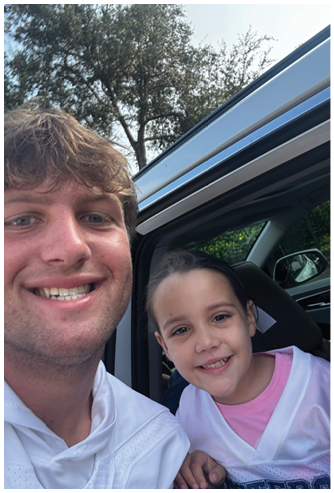 Two people smiling together near an open car door with trees visible in the background. Description generated by AI