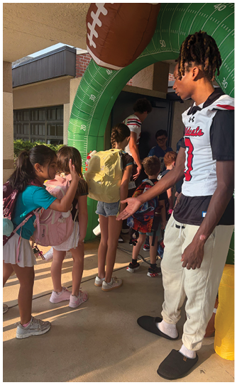 A group of people, including children with backpacks and an adult in a sports jersey, are gathered near a large green inflatable arch marked with measurement lines. Description generated by AI