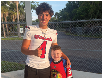 An adult wearing a Wildcats football jersey with the number 4 is giving a thumbs up and standing next to a child in a red sports jersey with the number 9, both smiling outdoors near a chain-link fence. Description generated by AI