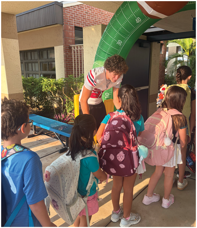 A group of children with backpacks are standing in line outside a building while an adult leans forward to speak to one of them under a large inflatable football goalpost. Description generated by AI