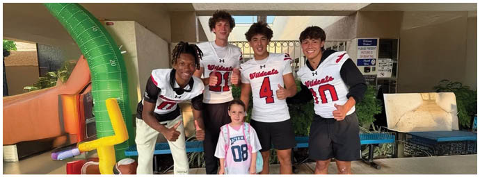 Five people wearing sports jerseys and one child with a backpack are posing and smiling together in an outdoor area near playground equipment. Description generated by AI