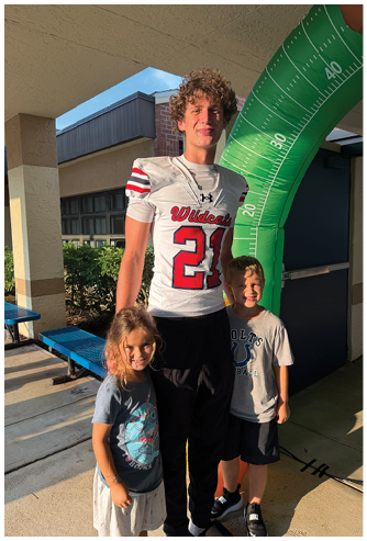 An adult wearing a football jersey stands smiling with two children in casual clothing near a green inflatable structure outside a building. Description generated by AI