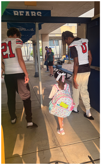 Two people in sports jerseys walk alongside a child with a backpack under a covered walkway near a school entrance. Description generated by AI