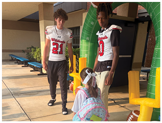 Three people wearing football jerseys stand on a sidewalk near inflatable football goalposts while one person with a backpack faces them. Description generated by AI
