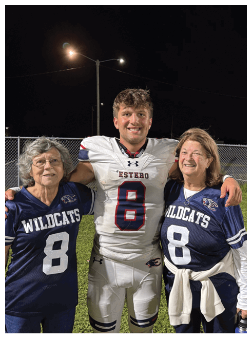 A person in a white football uniform with the number 8 stands smiling with arms around two people wearing blue Wildcats jerseys with the number 8 on a grassy field at night. Description generated by AI
