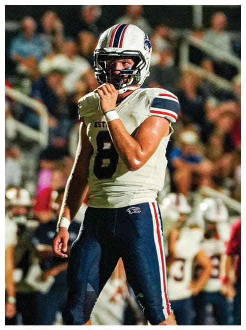 A person wearing a white football helmet with red and blue stripes and a white football jersey is holding their chin strap while looking ahead. Description generated by AI