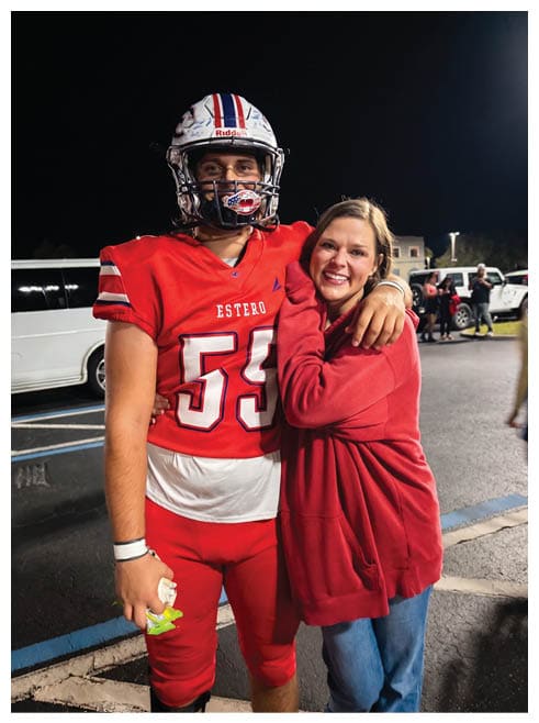 A person wearing a red football uniform and helmet stands next to another person in a red sweatshirt who is smiling and leaning on their shoulder against a dark background. Description generated by AI