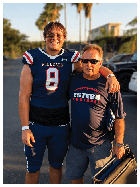 A person in a Wildcats football uniform stands with an arm around another person wearing sunglasses and an Estero Football shirt while holding a folded chair in a sunny outdoor parking area with palm trees in the background. Description generated by AI
