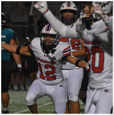 A person wearing a white football uniform with the number 12 and the word Wildcats is celebrating on a football field with arms outstretched. Description generated by AI