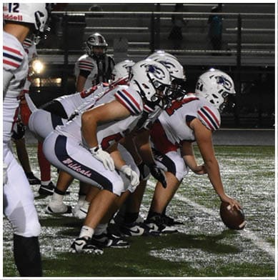 Players in football uniforms are lined up on the field preparing for a play with spectators seated sparsely in the bleachers behind them at night. Description generated by AI