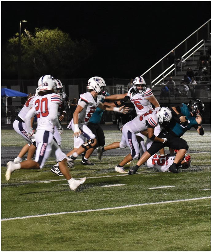 A group of people wearing football uniforms are actively engaged in a nighttime game on a field with spectators in the background. Description generated by AI