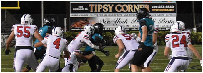 A group of people in football uniforms are engaged in a play on a field with a scoreboard in the background showing the score and time remaining. Description generated by AI