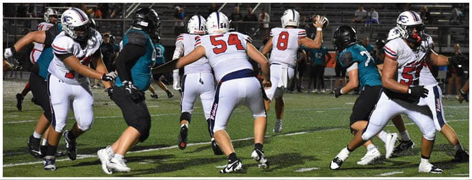 People wearing football uniforms are actively engaged in a game on a grassy field under stadium lights. Description generated by AI